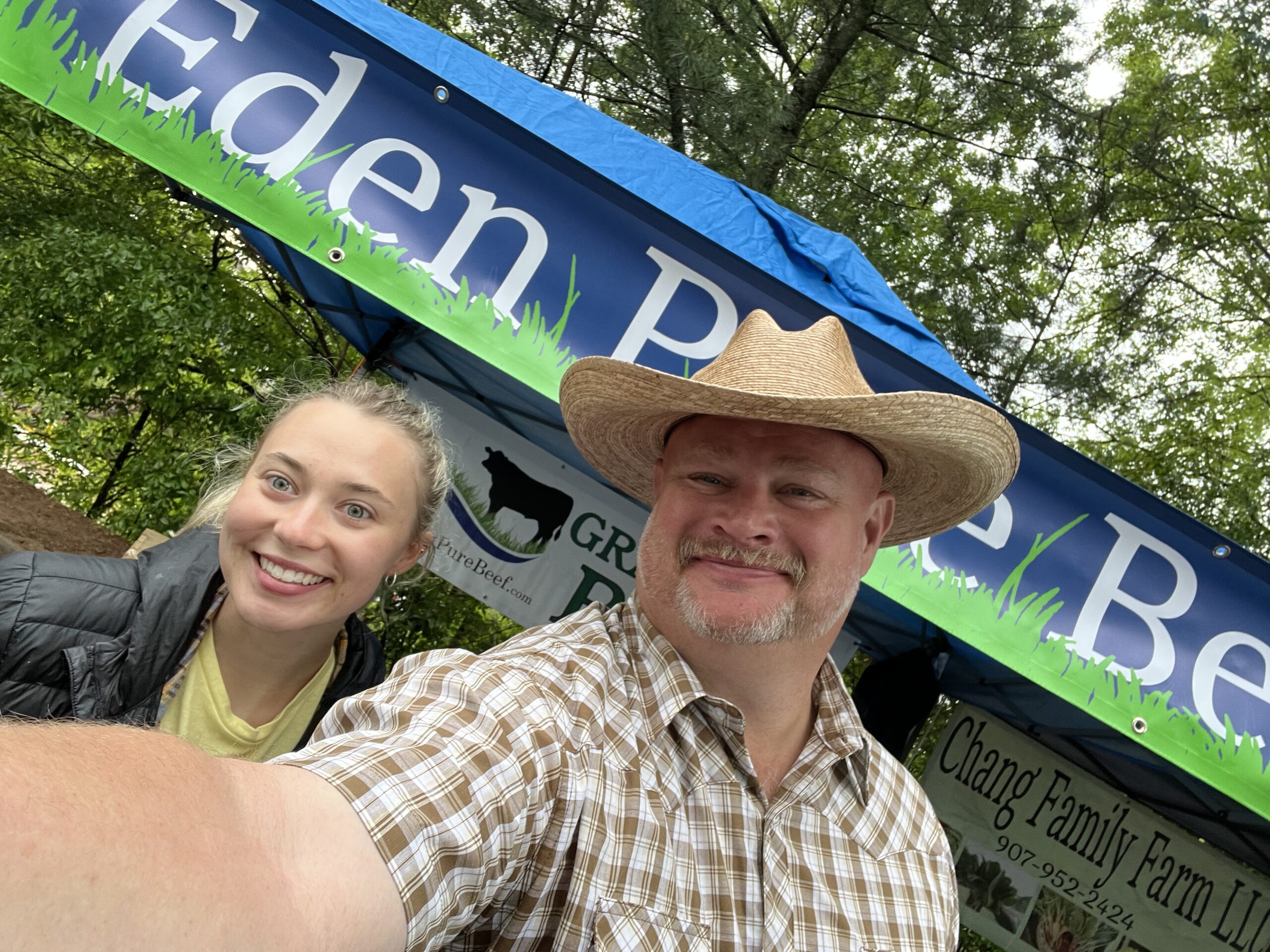 Farmer Emmet - Maraket BOoth 2 EPB Two smiling individuals pose for a selfie in front of a vendor booth at a farmers market, showcasing a sign for "Eden Pure Beef" and "Chang Family Farm LLC." The man wears a straw hat and a plaid shirt, while the woman has a casual jacket on. The background features greenery and a vibrant blue canopy.